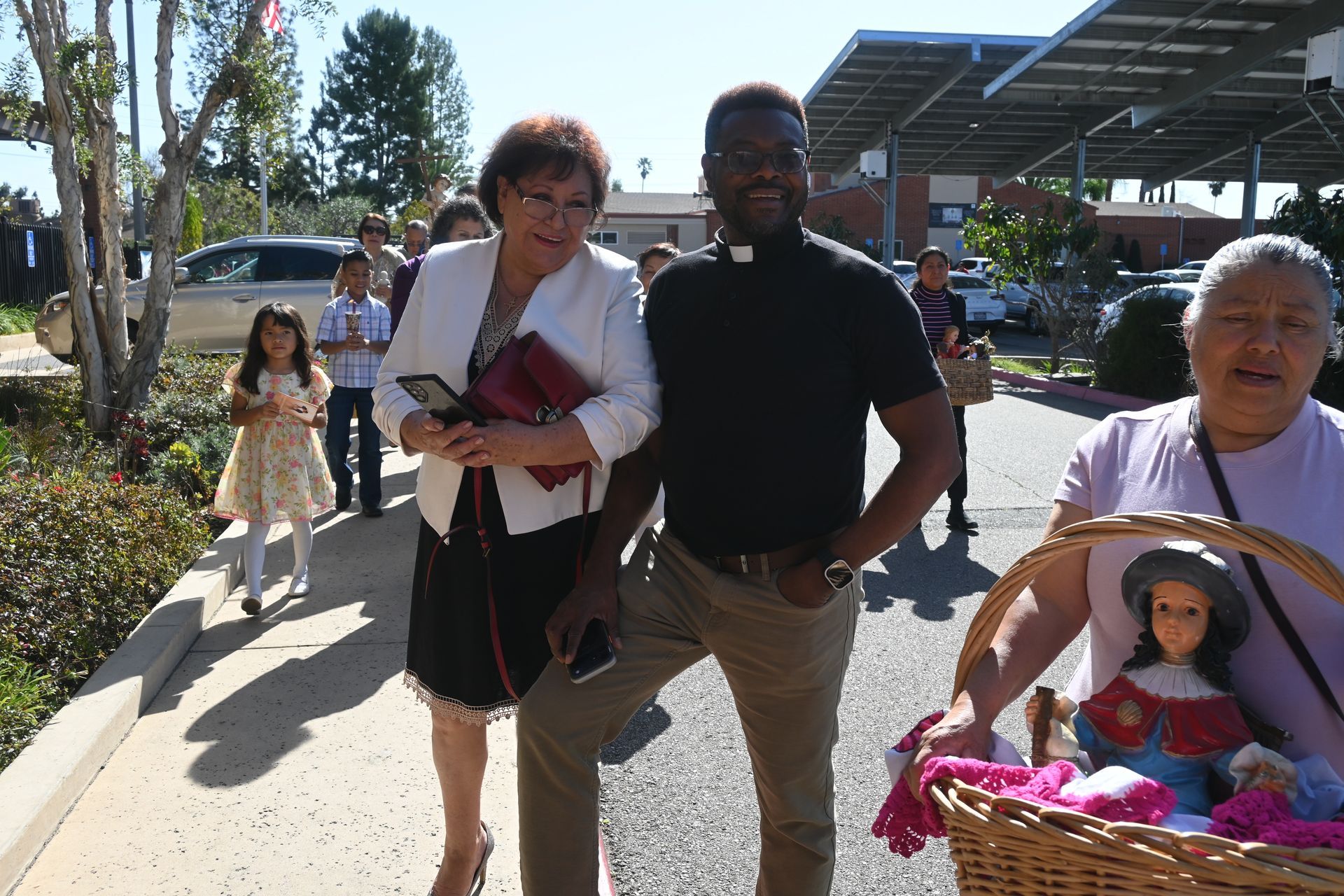 People walking outdoors on a sunny sidewalk, carrying bags and a basket with a doll.