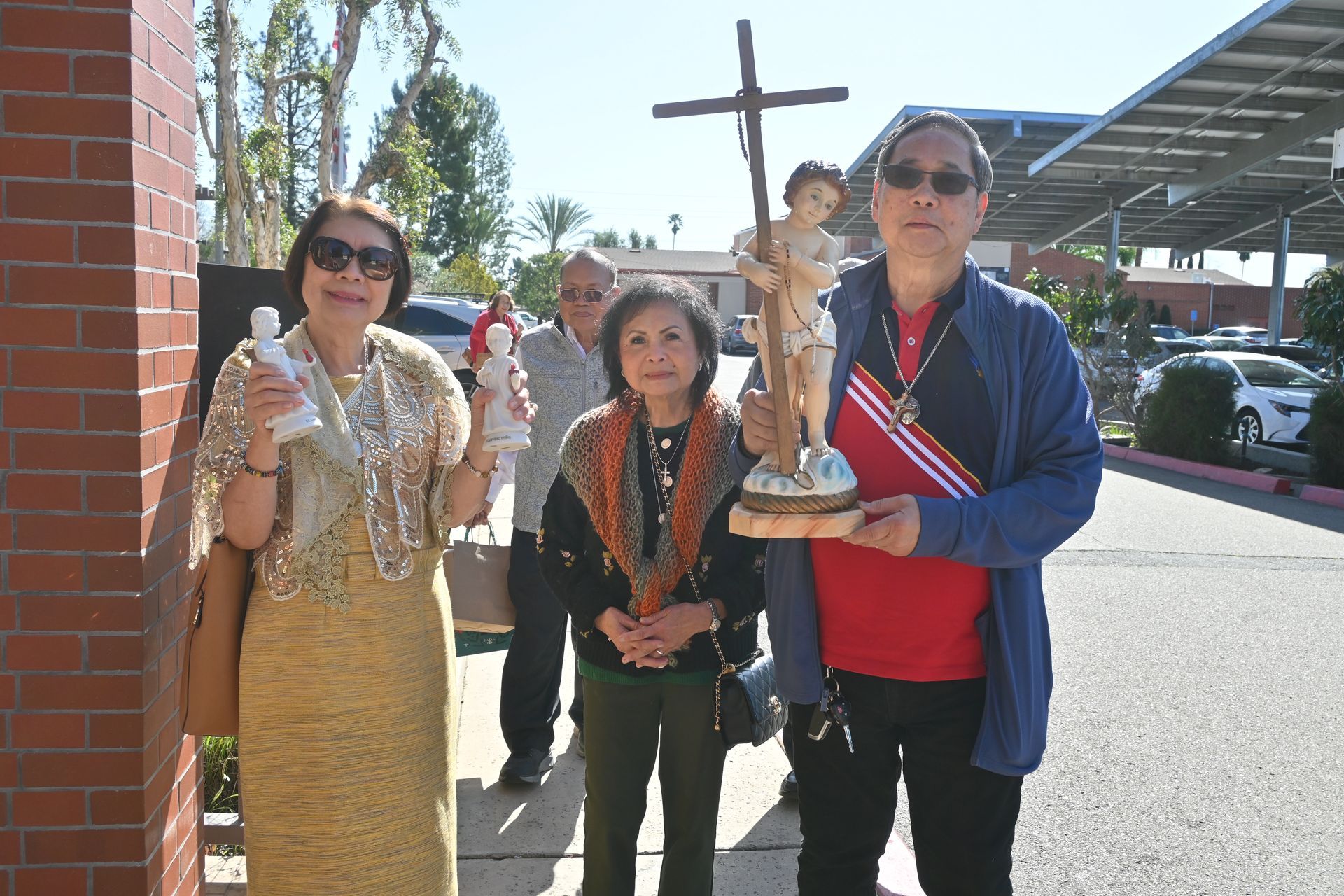 Three people holding decorative statues outside a brick building, with a cross in the background.