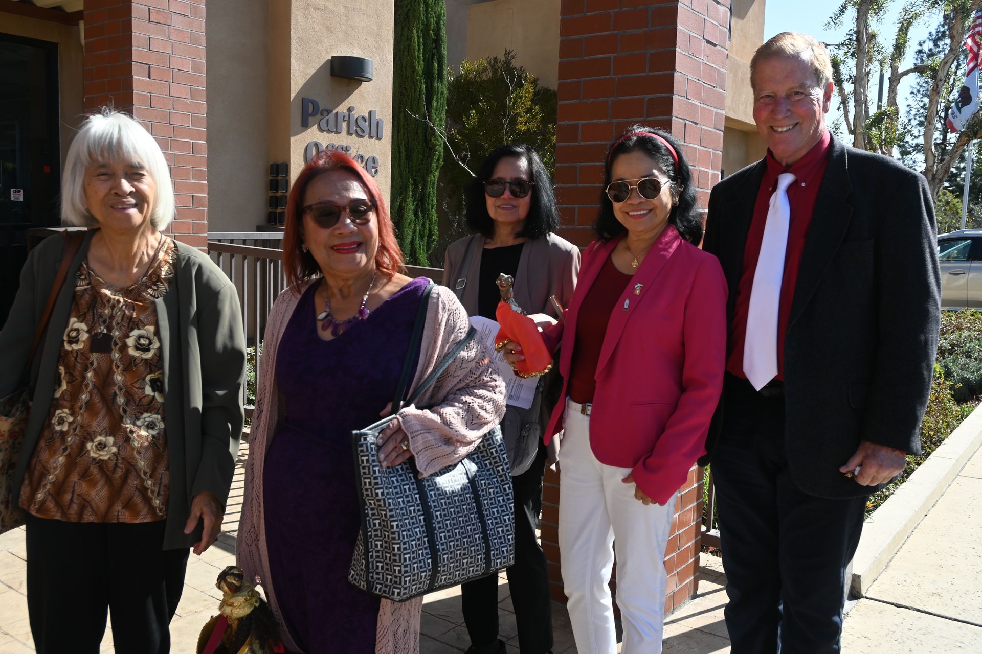Five people stand outdoors in bright sunlight, posing near a building entrance.