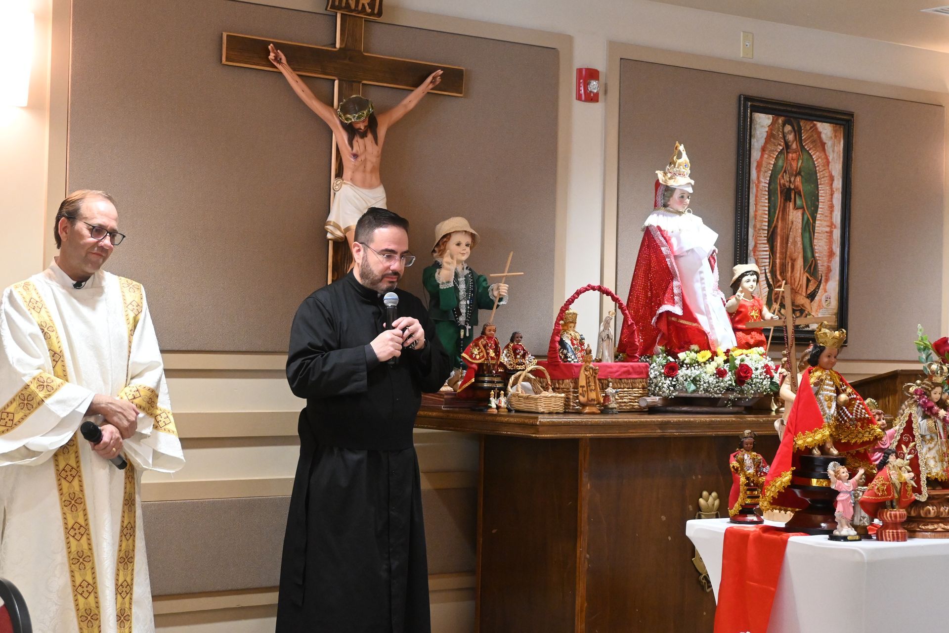 Priest speaking beside altar with crucifix and religious statues in a chapel