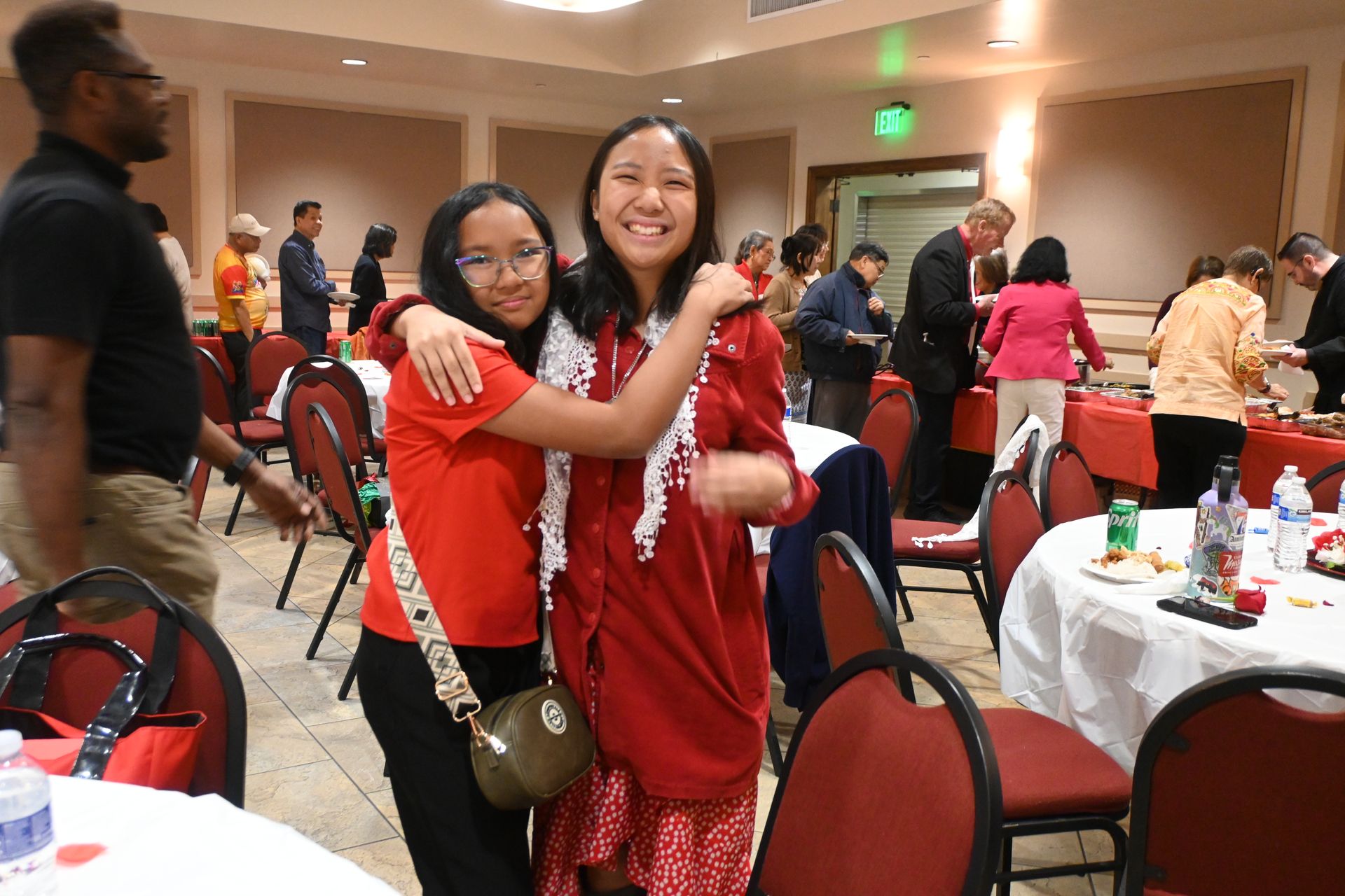 Two people in red dresses smiling and hugging at a banquet hall gathering.