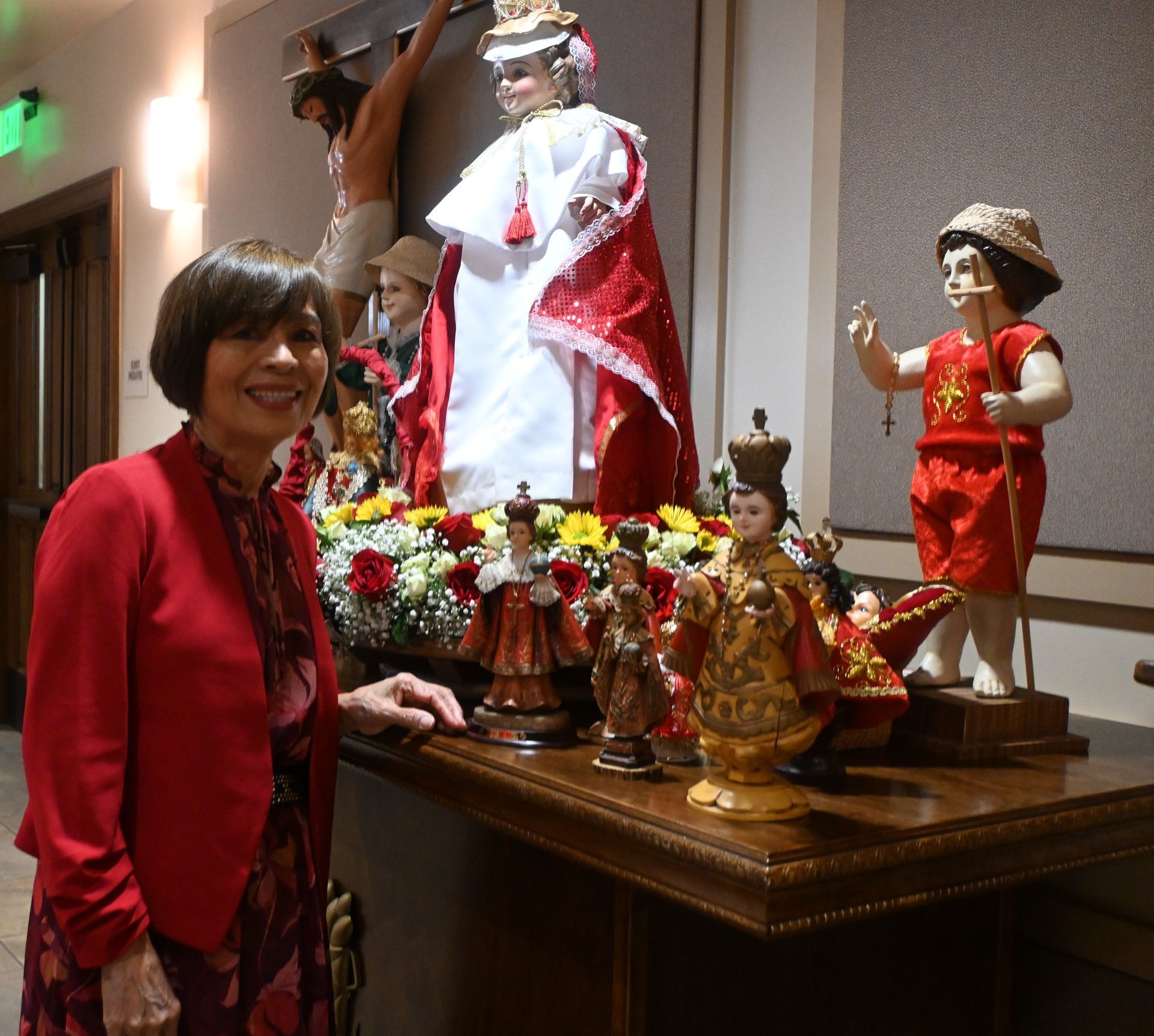 Woman beside a decorated altar with colorful figurines and flowers in a warmly lit indoor room