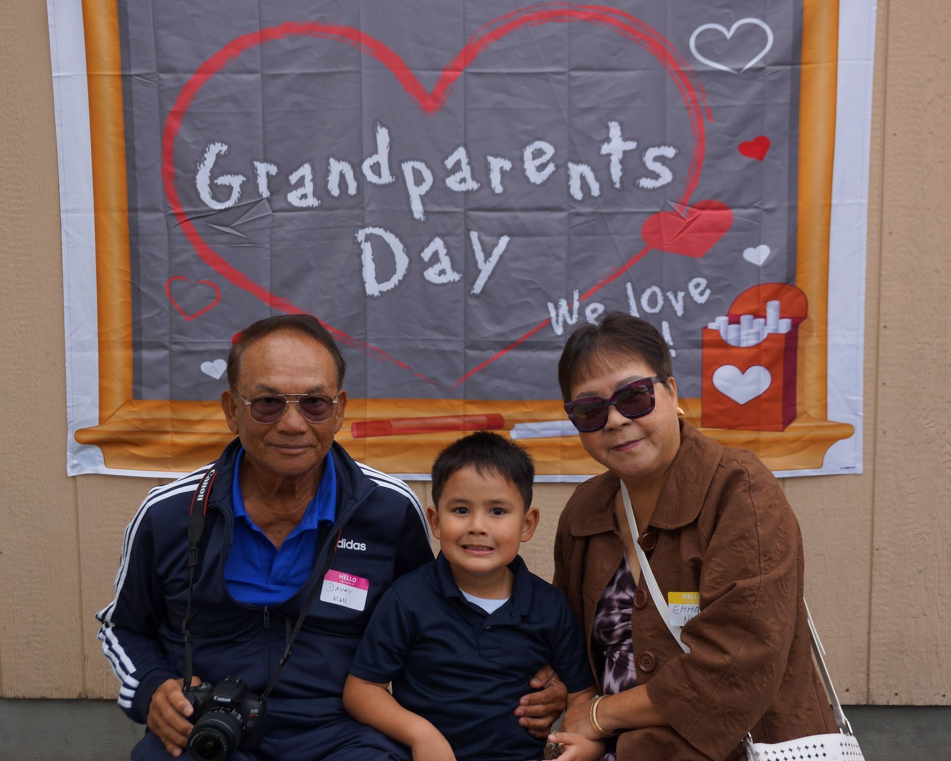 Grandparents Day photo with a smiling child between two seated adults in front of a chalkboard sign