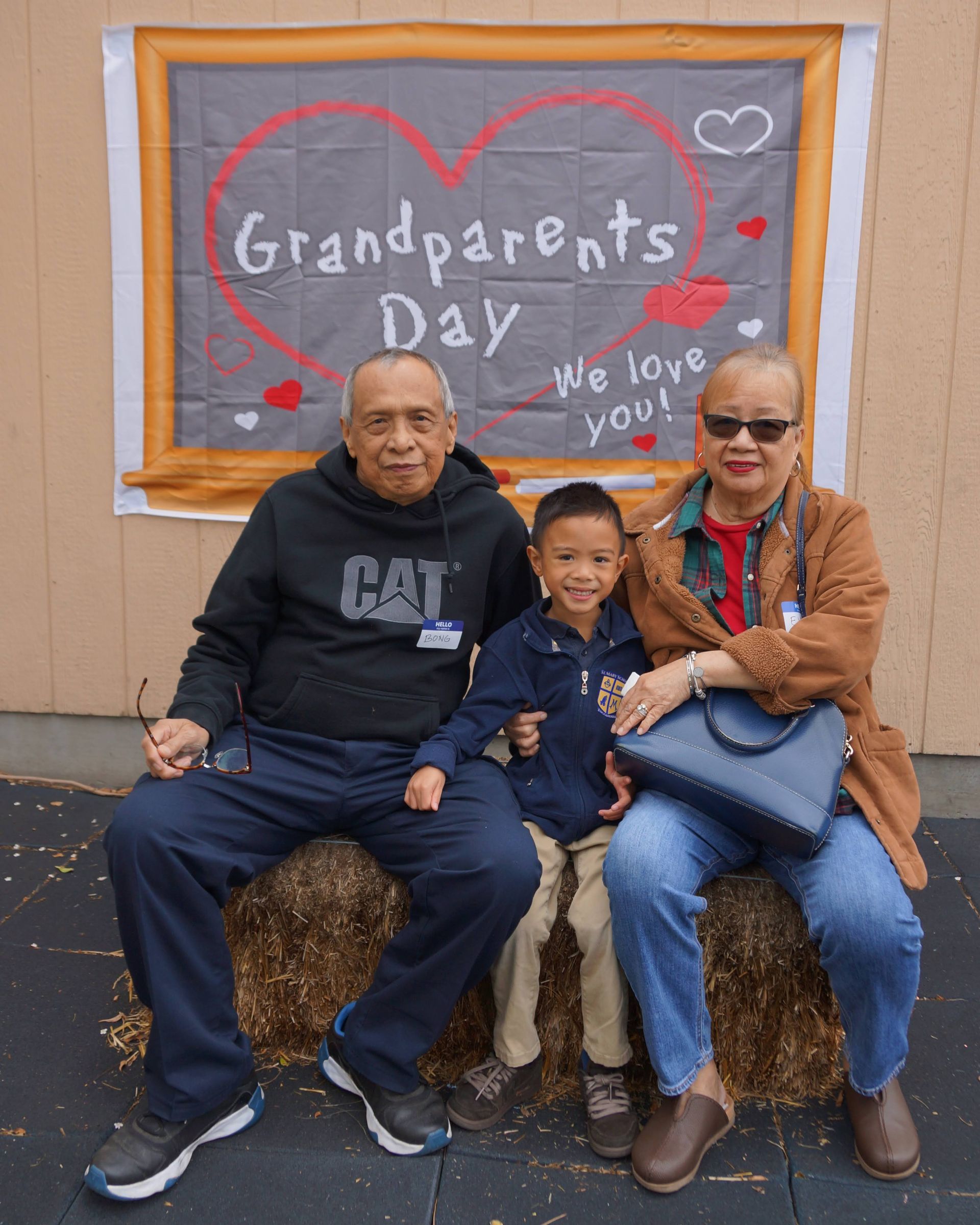 Three people seated in front of a chalkboard reading “Grandparents Day,” smiling for a photo.