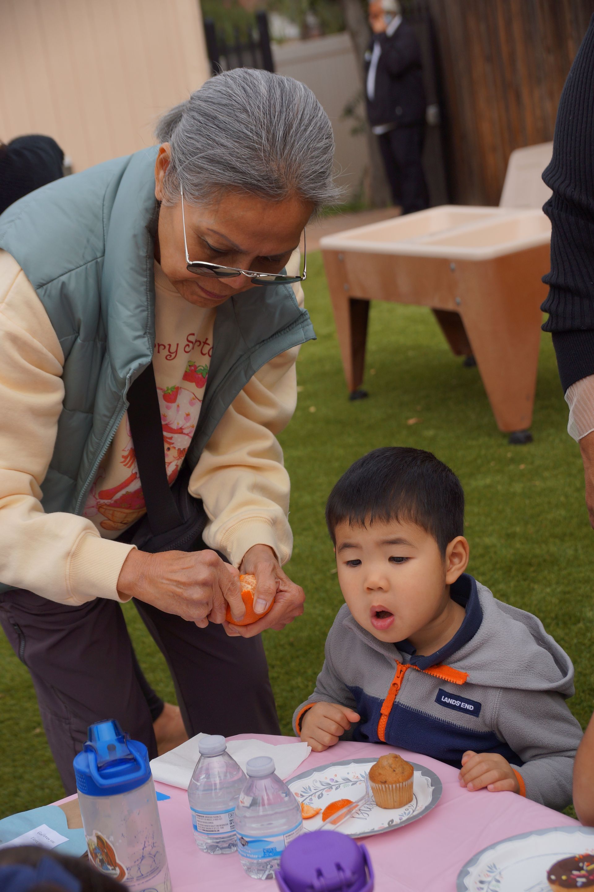 Adult serving food to a child at an outdoor table, both focused on a small plate and snack.