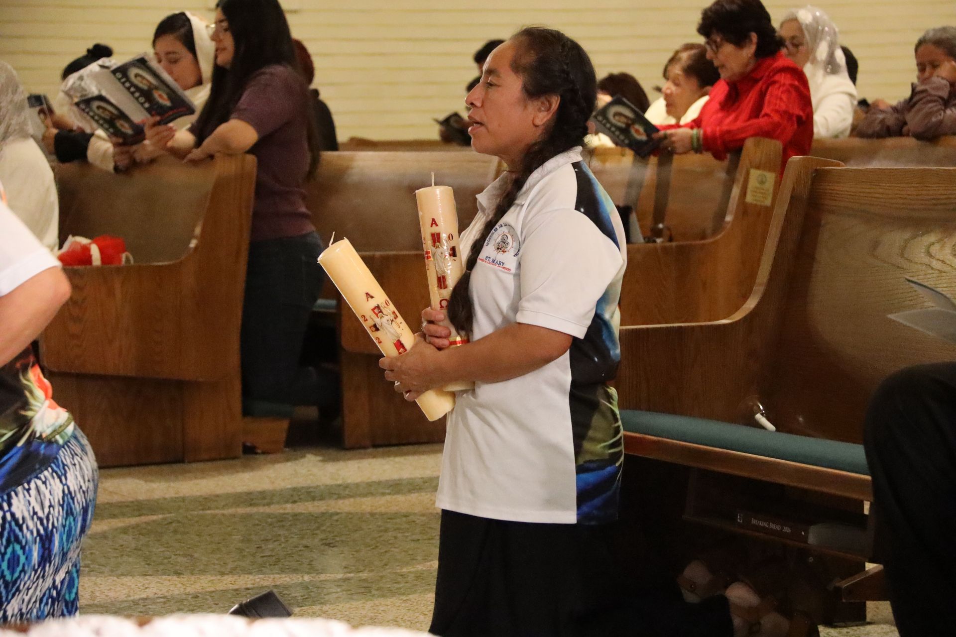 Person standing in a church pew holding two rolled papers, with seated people in the background