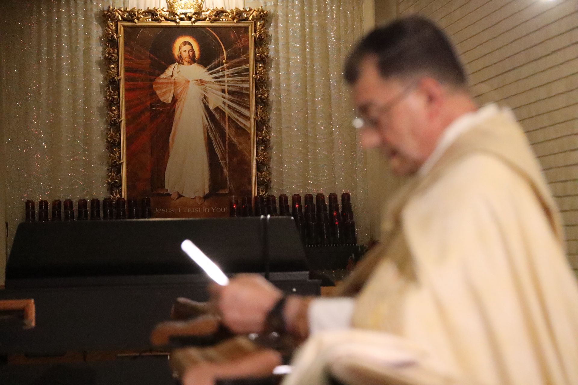 Priest in white vestments stands before a candlelit altar with a portrait of Jesus.