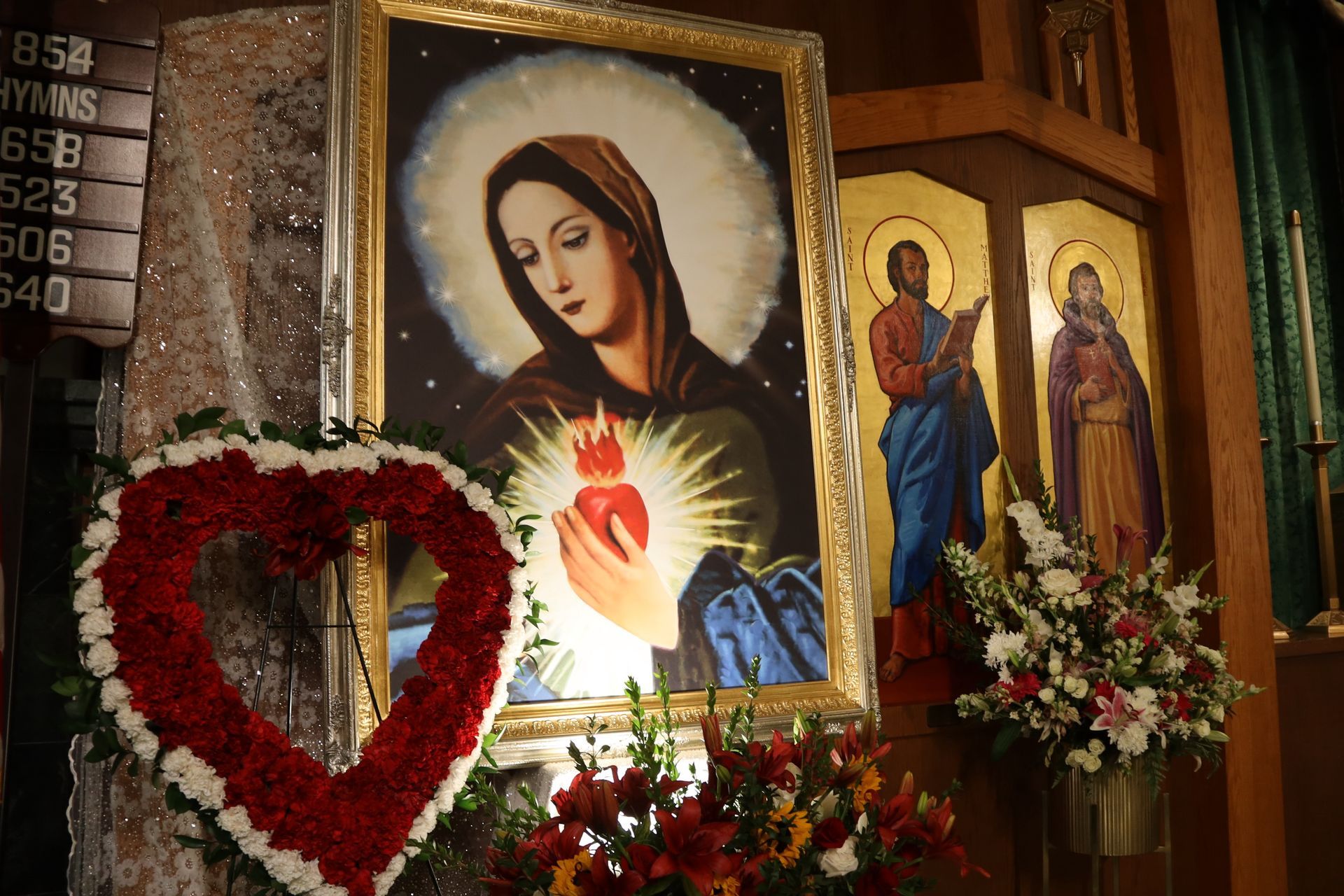 Religious altar with icons of the Virgin Mary and saints, red heart wreaths, and flowers in a church nook