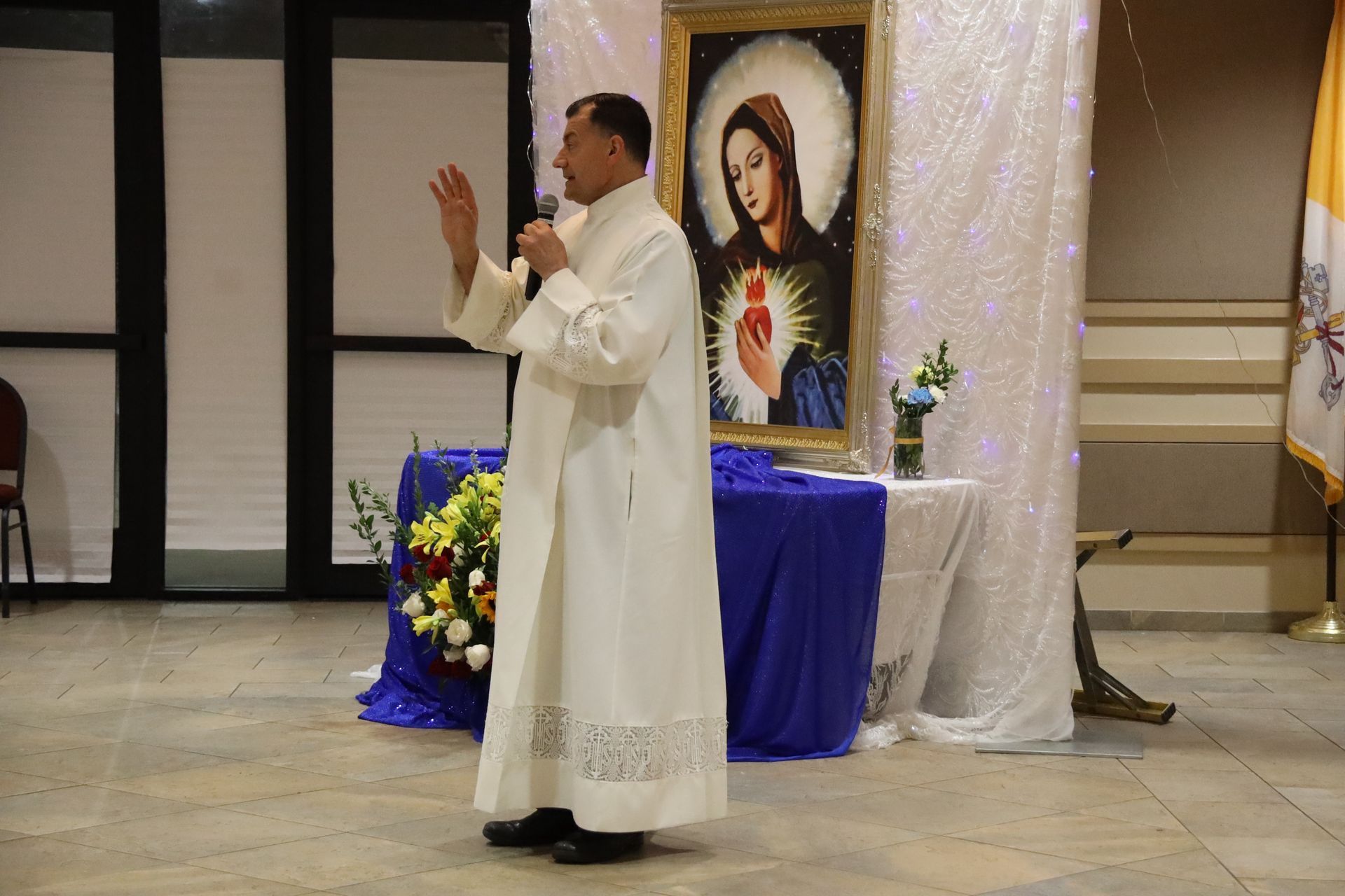 Priest in white robe speaking at a church altar beside a portrait of Jesus and flowers