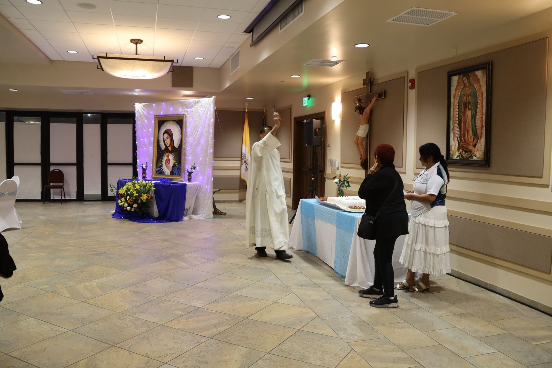 People gathered in a hall beside a white-draped altar with a framed religious icon and floral arrangements.