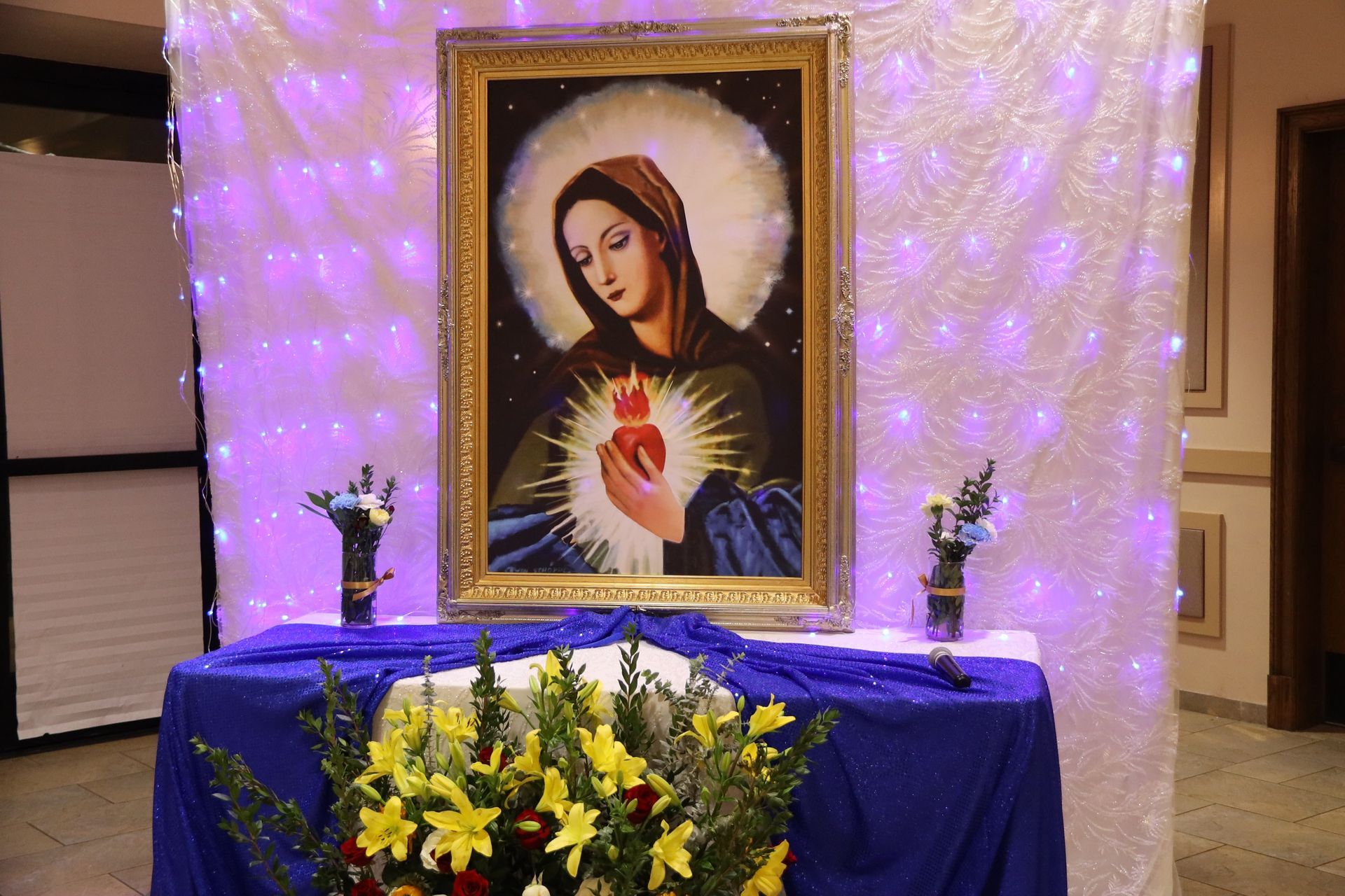 Religious altar with framed Virgin Mary portrait, blue cloth, flowers, and purple lights.