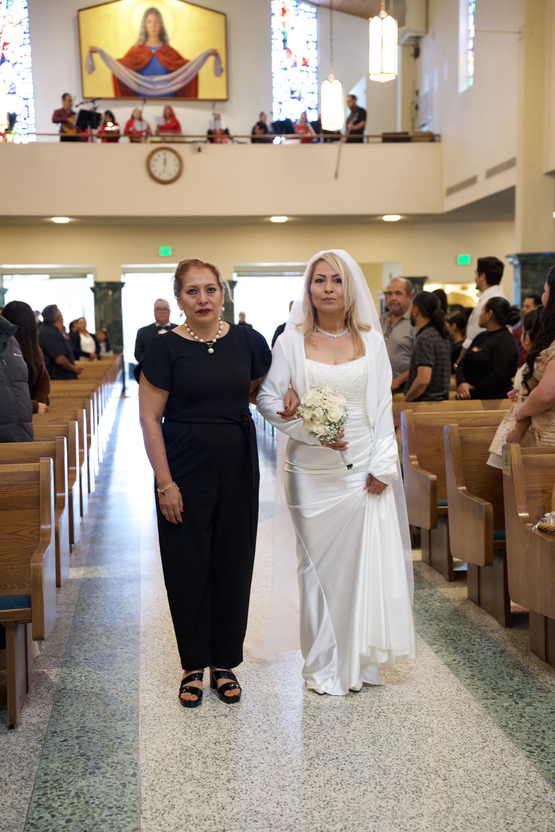 Two women in a church aisle, one in a white dress holding flowers, with pews and altar behind them.