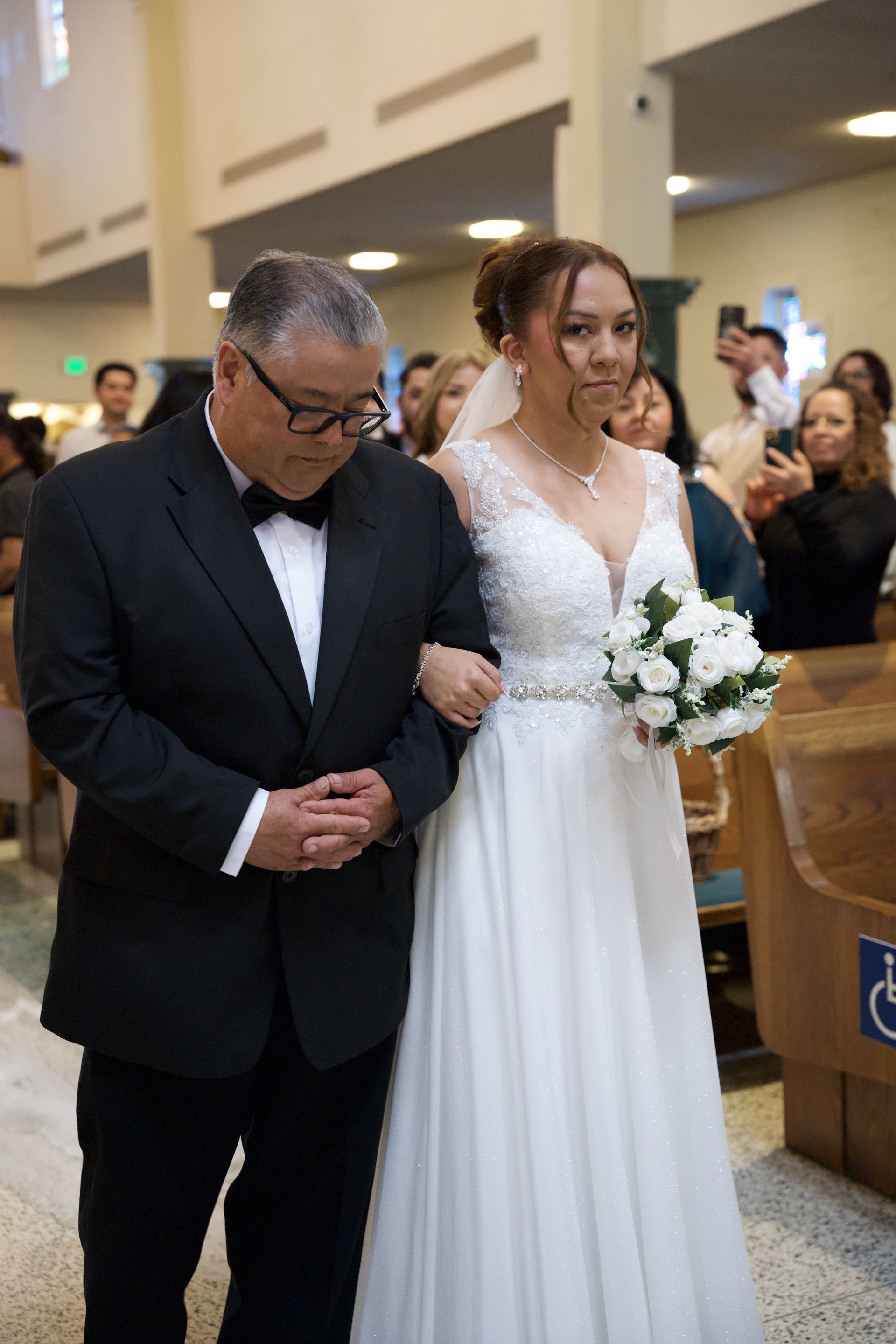 Bride in white dress walking arm-in-arm with a man in a black suit at a wedding ceremony.
