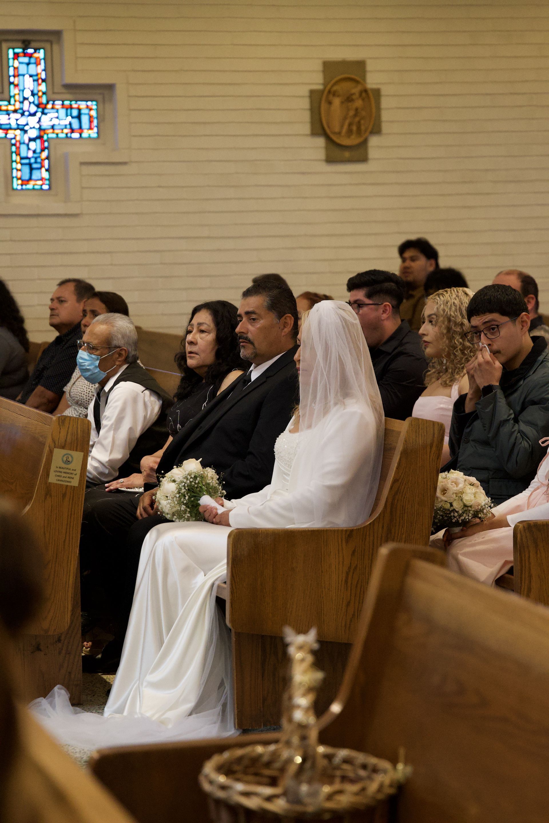 Bride in white veil seated in church pew during wedding ceremony, with guests nearby and stained glass in background