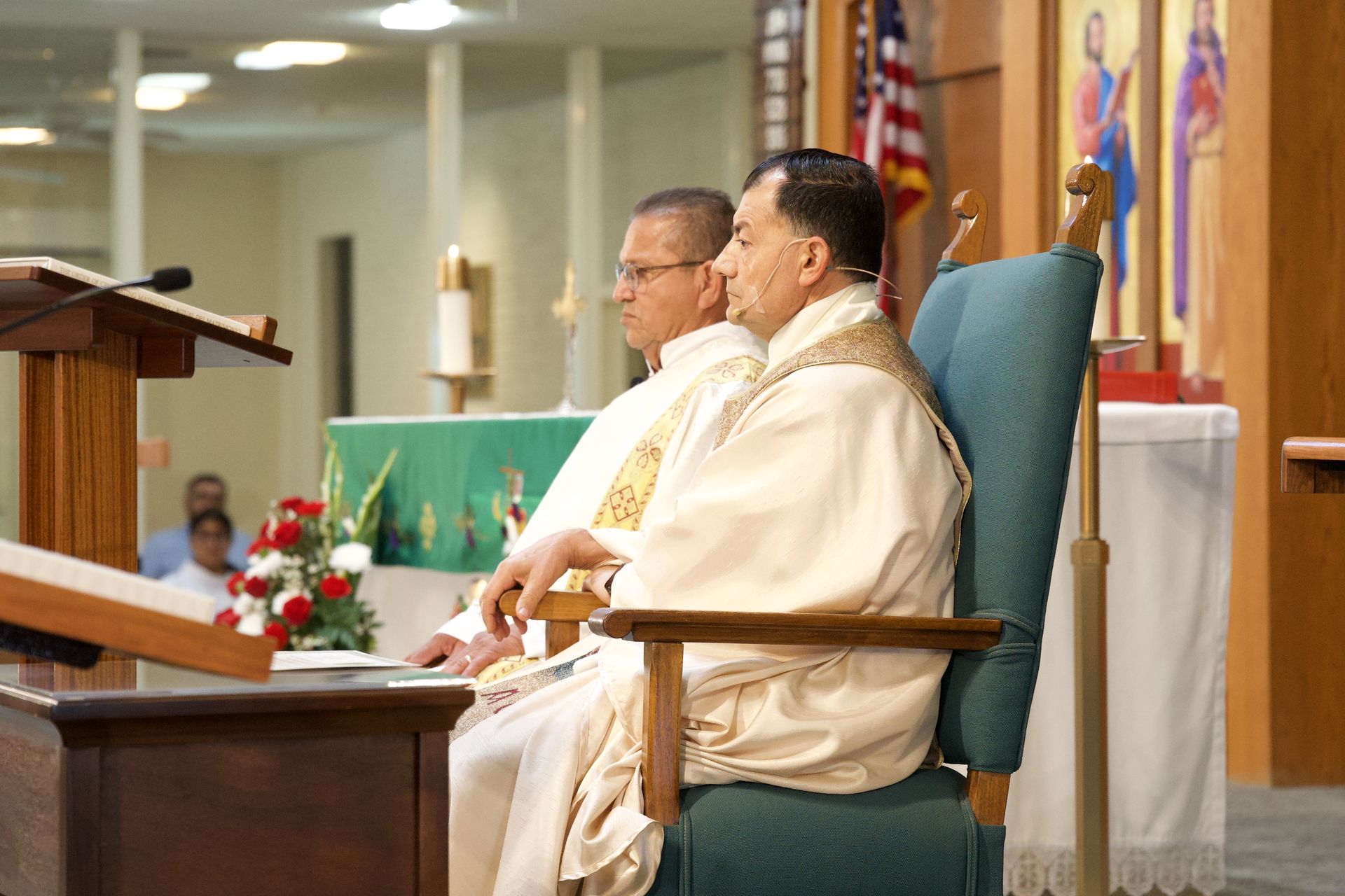 Two clergy members seated in a church beside a podium, facing the congregation.