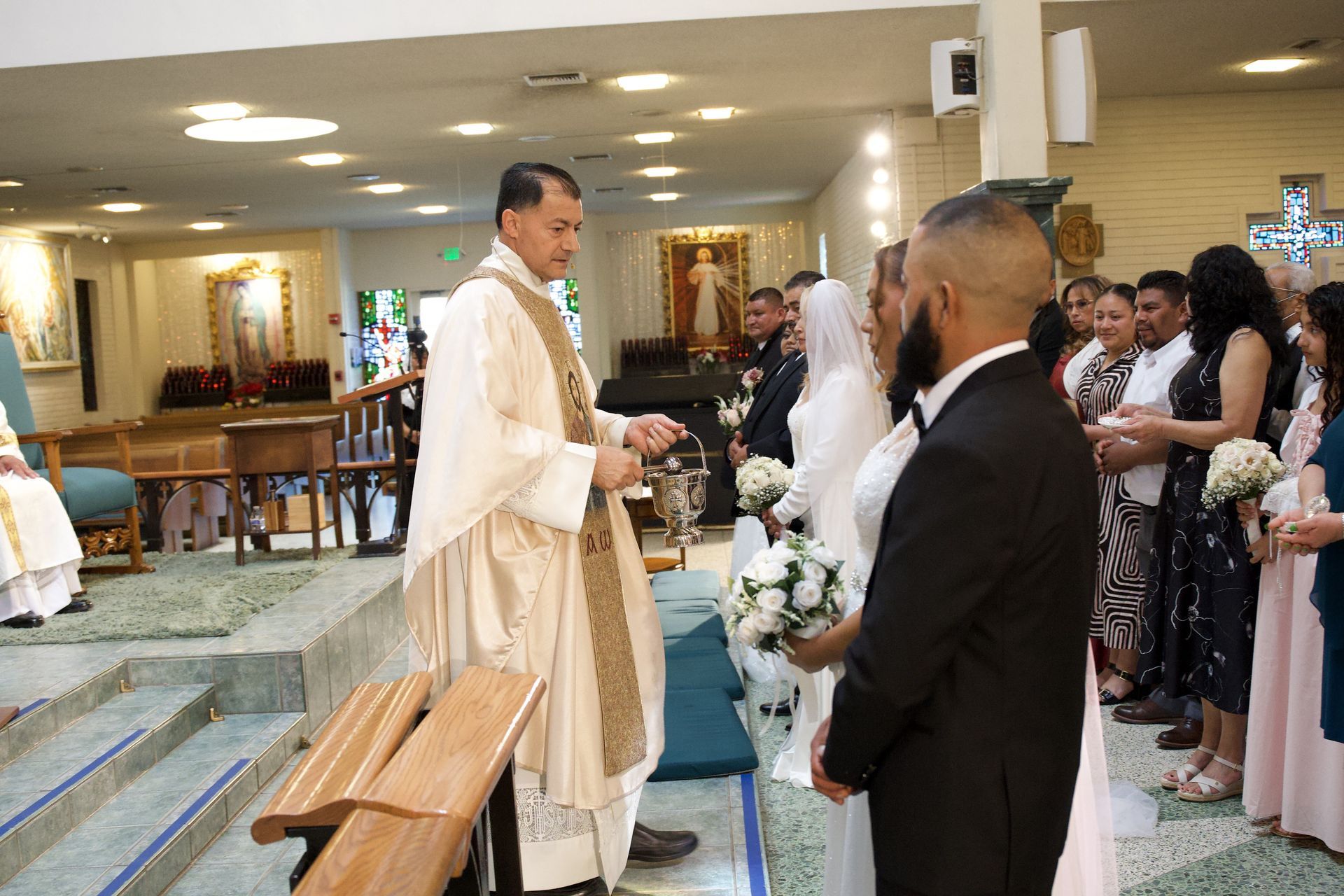 Priest in white and gold vestments greets guests in a church during a wedding ceremony