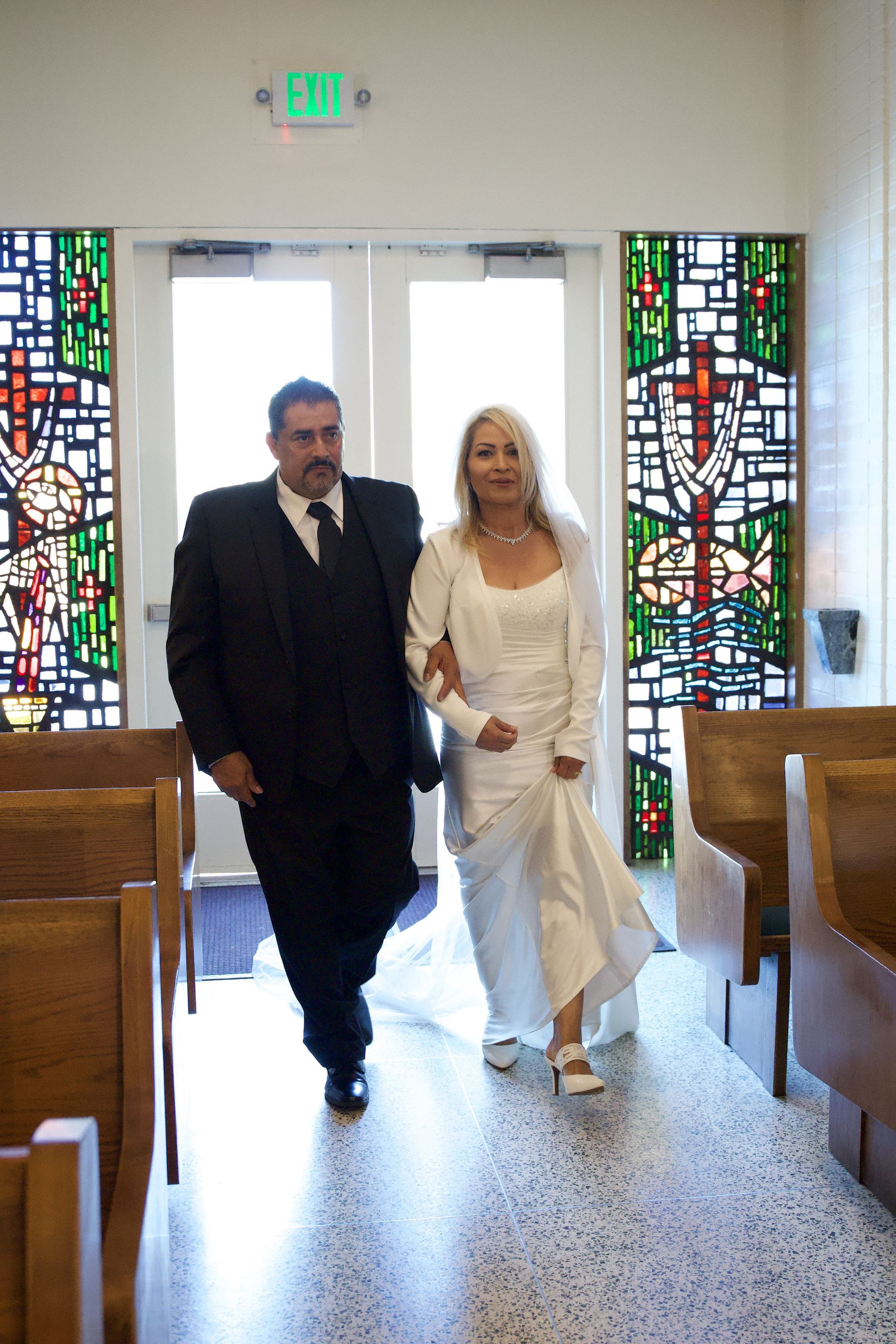 Couple walking down a church aisle, dressed in formal wedding attire, with stained-glass windows behind them.