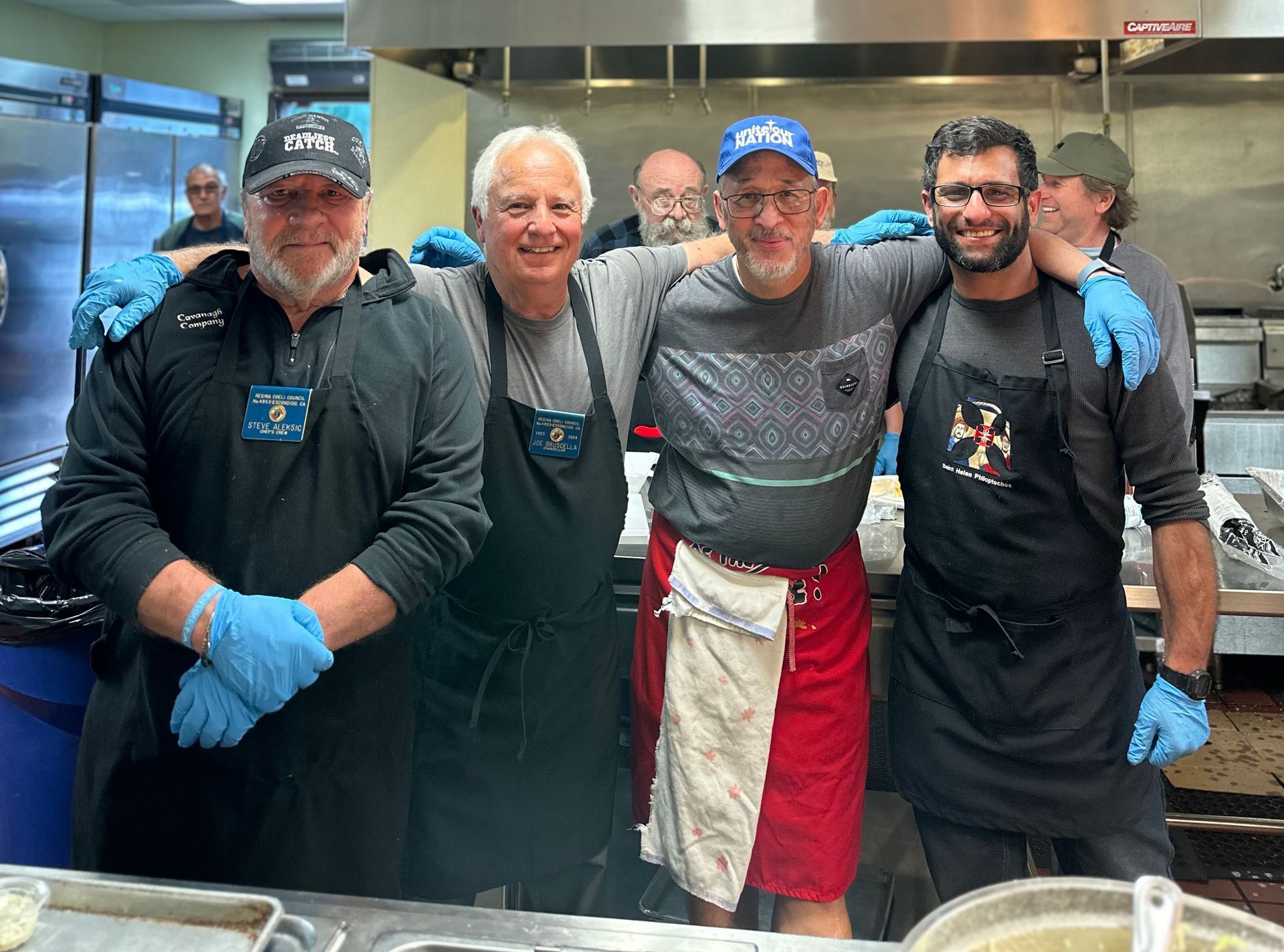 Four smiling kitchen staff in aprons and gloves stand together in a stainless-steel kitchen.