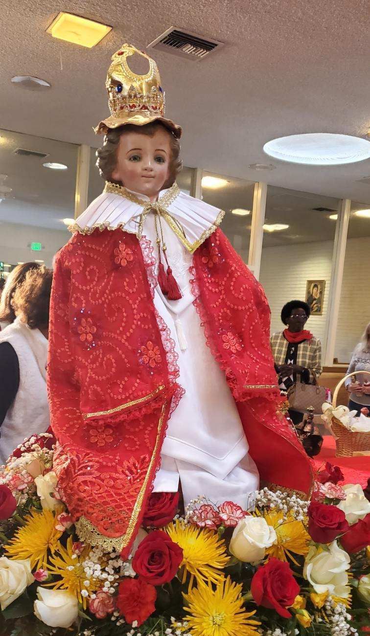 Decorated doll in red cape and white gown, standing among red and yellow flowers indoors