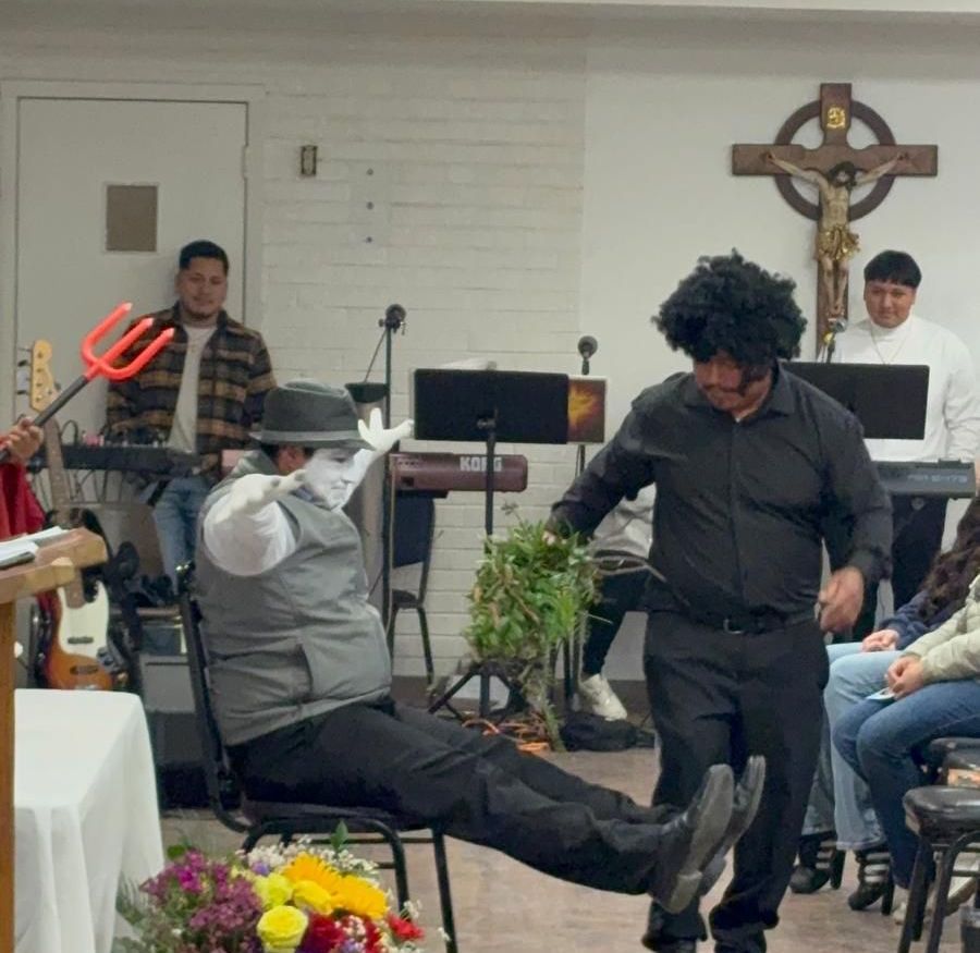 Man dancing at the front of a small church gathering, with people seated and standing nearby.