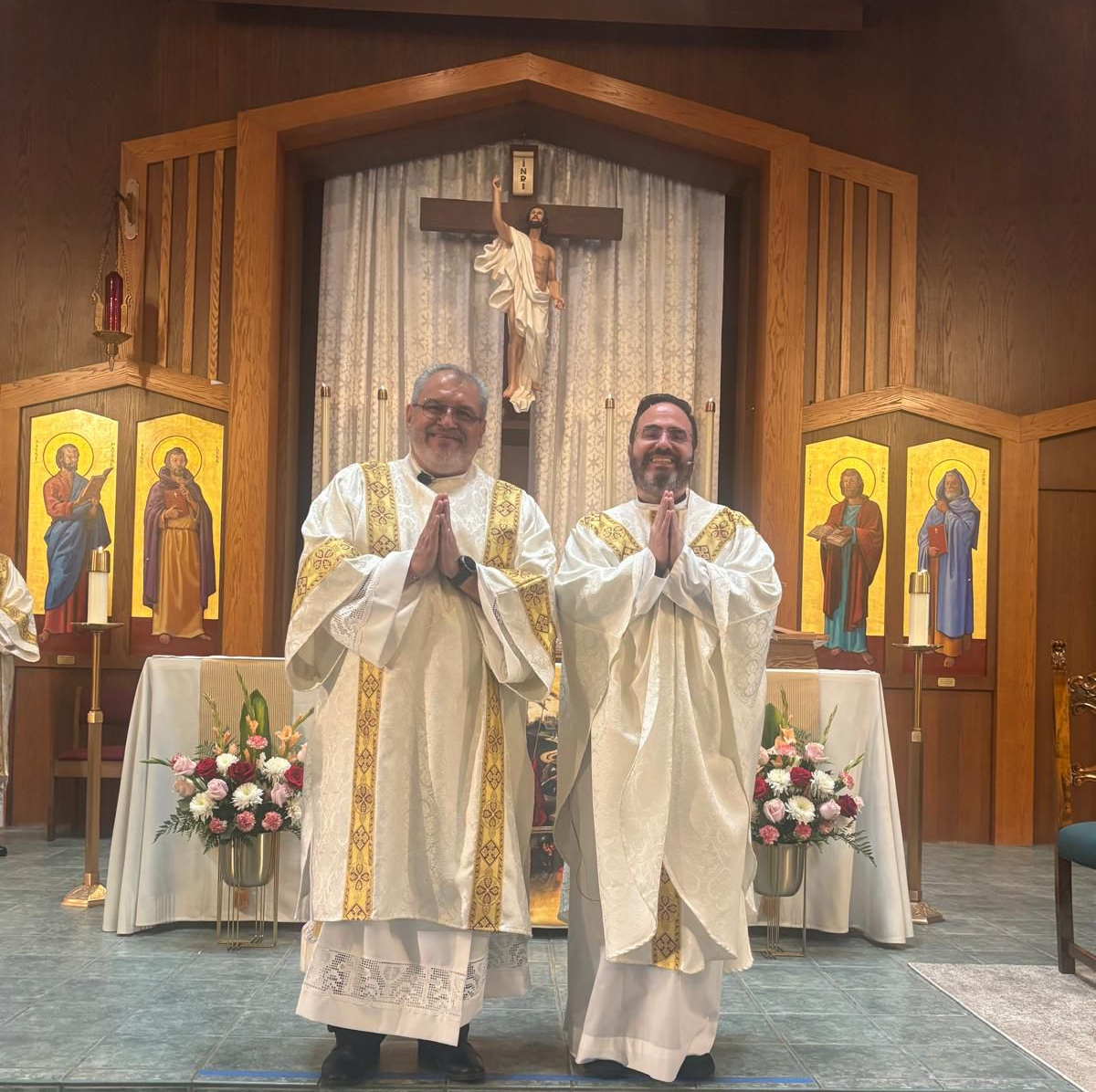 Two priests in white vestments stand smiling before an altar with a crucifix in a church.