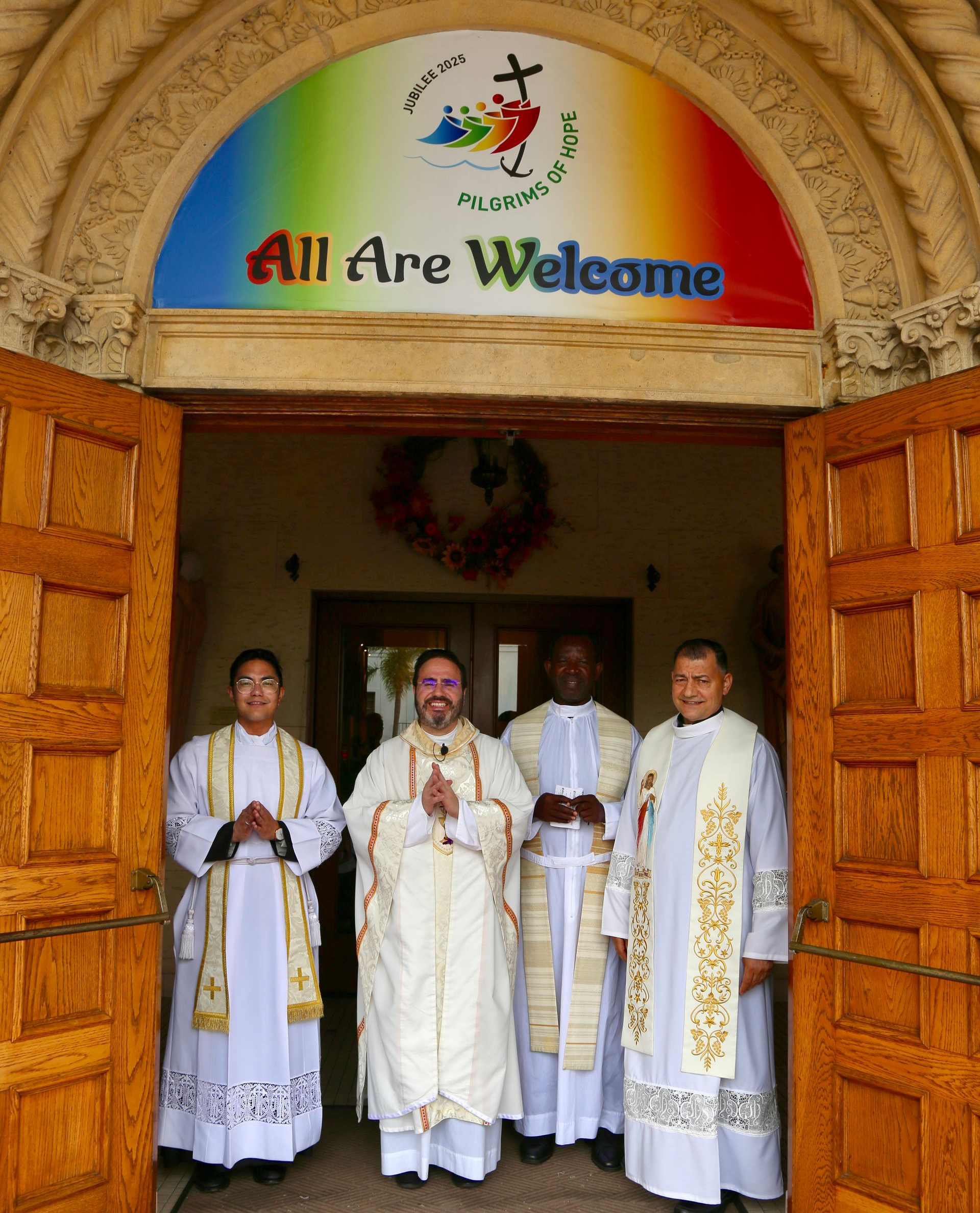 Four clergy in white vestments stand at a church doorway beneath an “All Are Welcome” sign.