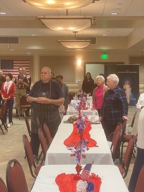 People standing around decorated tables in a banquet hall with red, white, and blue decorations.