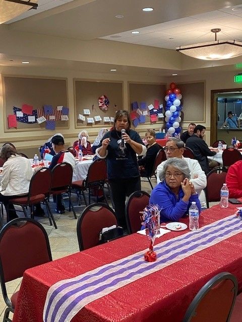 Banquet hall with people seated at decorated tables; balloons and red, white, and blue tablecloths on display.