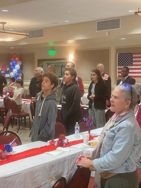 People standing attentively in a decorated banquet hall with patriotic red, white, and blue decor.