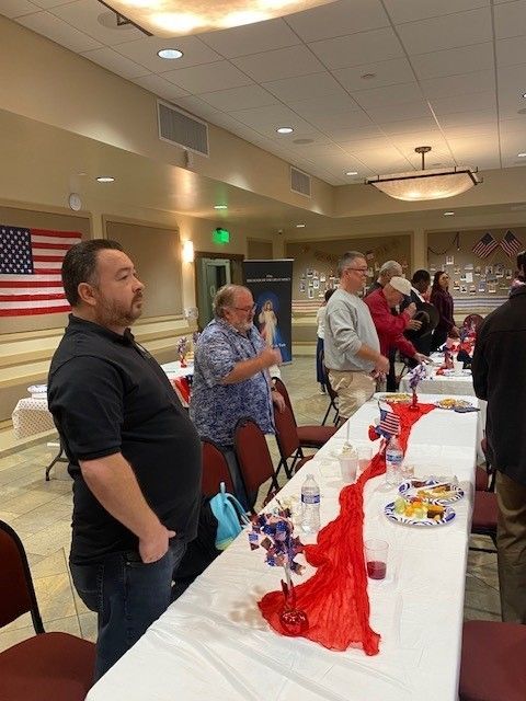 People standing by a decorated banquet table in a community hall with an American flag on the wall.