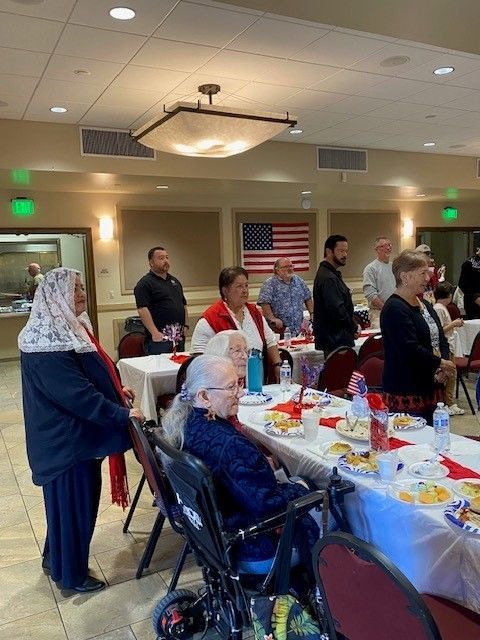 Community hall banquet with people standing around a decorated table under warm ceiling lights.