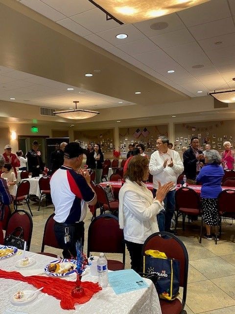 People standing and applauding at a banquet hall event with red tablecloths and round tables