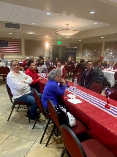 People seated at banquet tables in a community hall, facing a speaker or event off-camera.