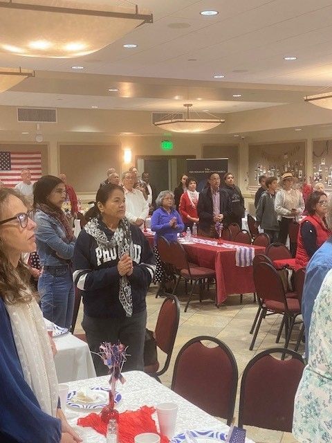 People standing in a banquet hall during a gathering, with tables, chairs, and an American flag in the background.