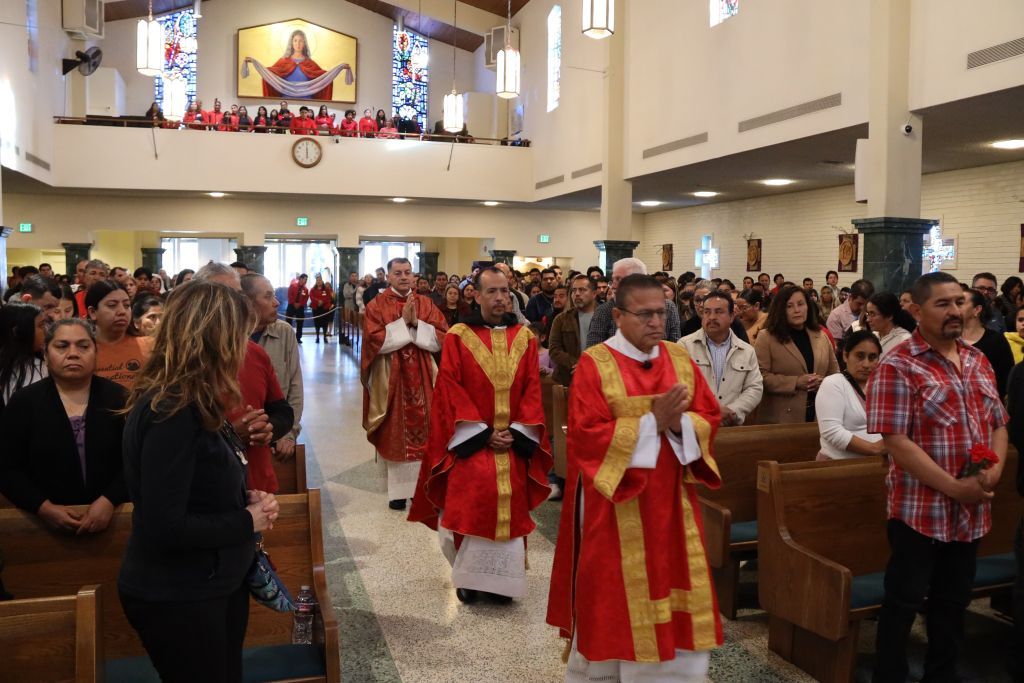 Clergy in red and gold robes process down a church aisle during a crowded ceremony.