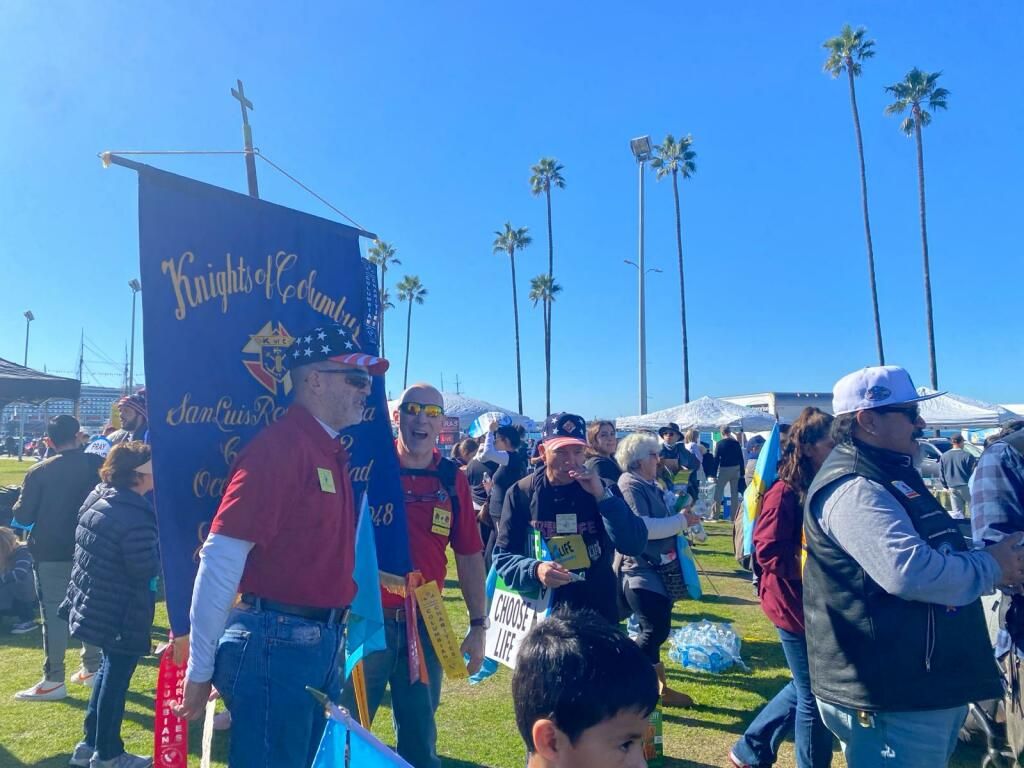 Crowd at a sunny outdoor event with a blue sign, palm trees, and people wearing jackets and hats.