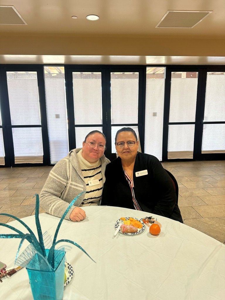 Two people seated at a round table in a bright lobby, smiling with drinks and snacks nearby