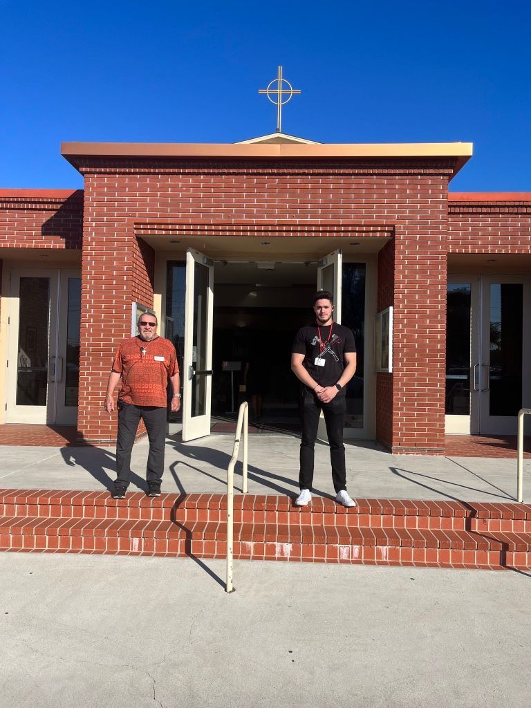 Two people stand at the entrance of a brick church with a cross on top.