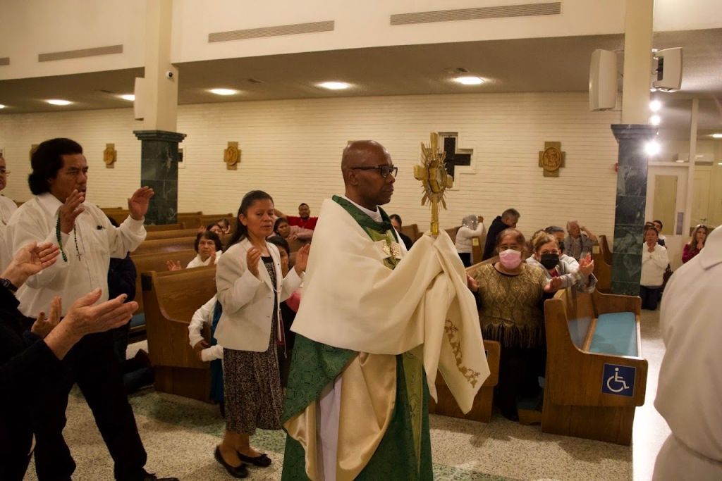 Priest in white and green vestments leading a church procession with congregants clapping nearby