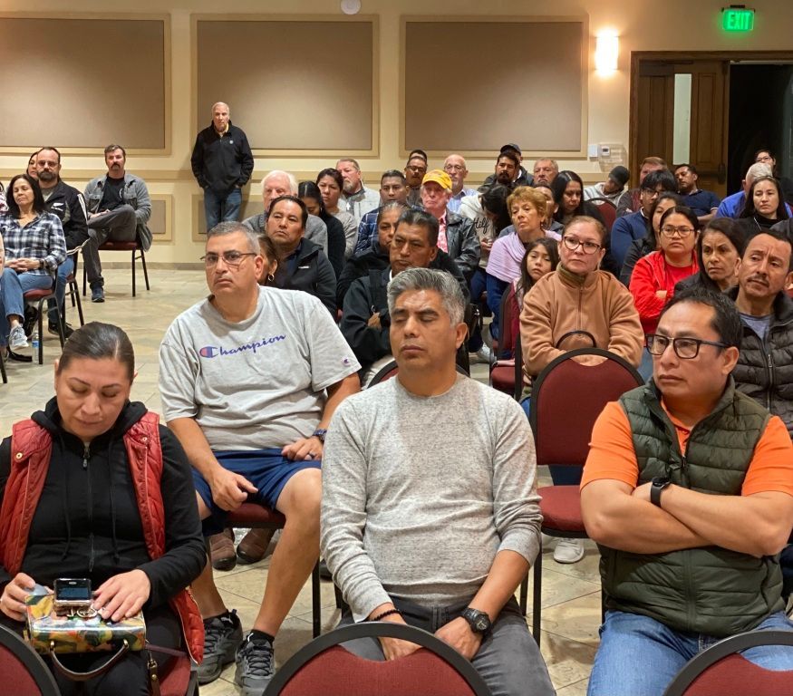 Seated audience in a meeting room, facing forward and listening attentively.