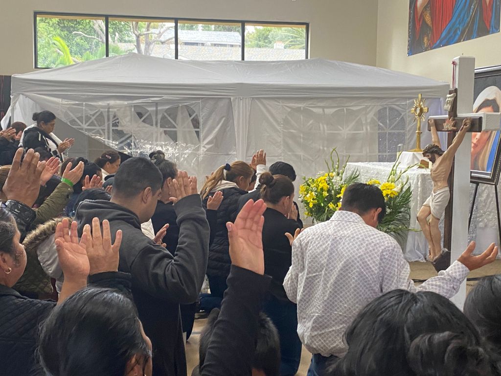 Crowd worshipping with raised hands in a bright indoor tent beside a cross and floral arrangement