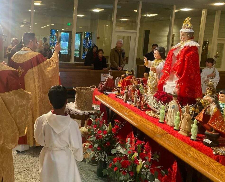 Person in white robes faces a decorated saint statue in a church, with candles and flowers nearby