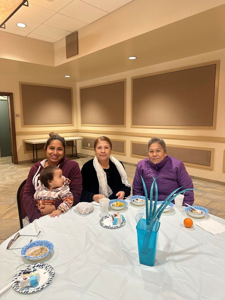 Three people sit at a table in a banquet room with a baby, blue cups, and plates.