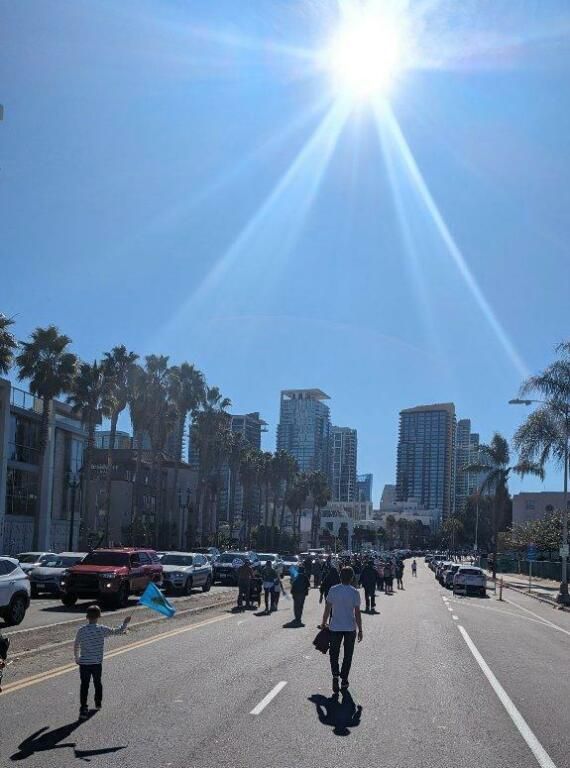 People walking along a sunlit city street lined with palm trees and tall buildings.