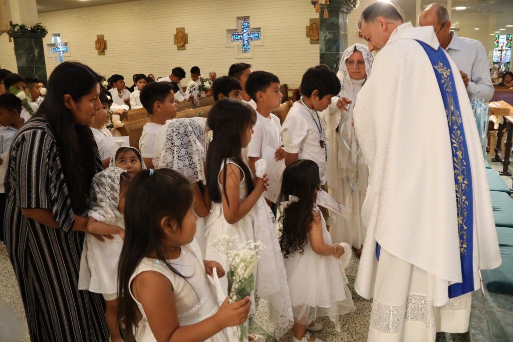 Children in white dresses receiving communion from a priest in a church sanctuary