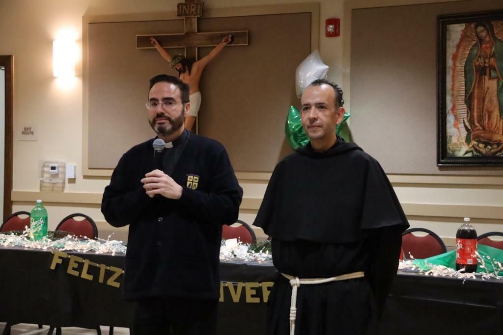 Two men stand indoors before a crucifix and altar, one speaking into a microphone.