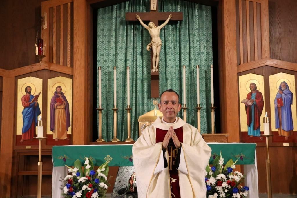 Priest praying at altar beneath crucifix, with green backdrop, candles, and religious icons on either side