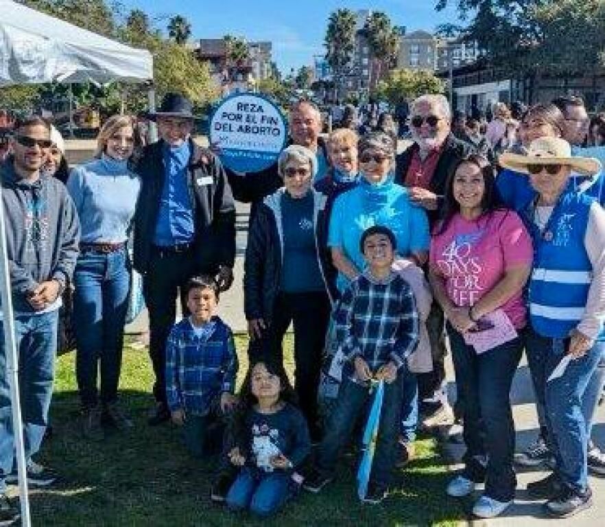 Group of people posing outdoors in blue shirts and hats at a community event, with a sign in front.