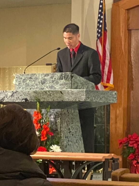 Man speaking at a marble podium in a church, with an American flag and red flowers nearby.