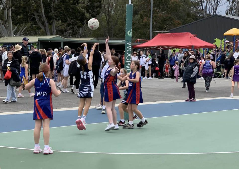 Girls play netball on an outdoor court with a crowd and red tents in the background.